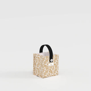 A small square structured gift bag with an all-over gold botanical print on a cream background, featuring a black webbing handle attached with a silver grommet.
