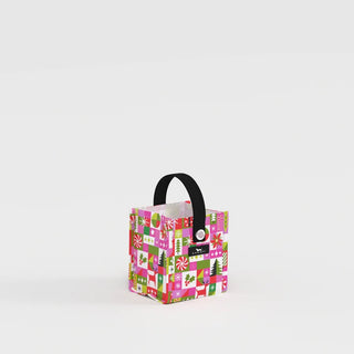 A small square structured gift bag in a multicolor holiday patchwork print featuring trees, reindeer, holly, and snowflakes in pink, green, red, and lavender, with a short black webbing handle and metal grommet.