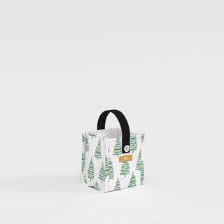 A small structured square gift bag with a white background printed with watercolor green evergreen trees and gold accent dots, featuring a black woven webbing handle and a metal grommet, shown from a slight angle.
