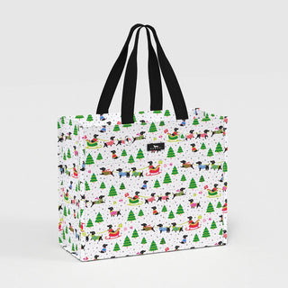 A structured large tote-style gift bag with black woven handles, white exterior printed with black dachshund dogs in colorful holiday sweaters, green evergreen trees, red sleds, and gray polka dots.