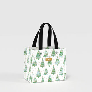 A small structured rectangular gift bag with a white ground covered in watercolor-style green evergreen trees with gold star and ornament accents, black woven webbing handles, silver grommet hardware, and a gold logo label plate on the front.