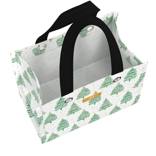 A small structured rectangular gift bag with a white ground covered in watercolor-style green evergreen trees with gold star and ornament accents, black woven webbing handles, silver grommet hardware, and a gold logo label plate on the front.