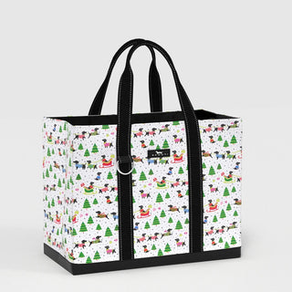 A structured large rectangular tote bag with a white ground printed with dachshunds in colorful sweaters, green holiday trees, sleighs, and gift boxes, with dual black webbing handles and a black reinforced base.