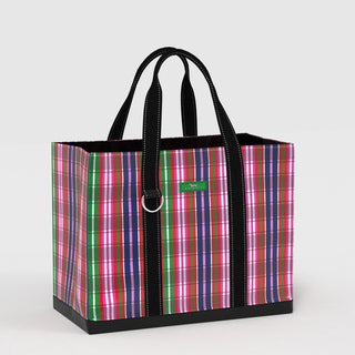 A large structured open-top tote bag in a multicolor plaid print of pink, green, navy, red, lavender, and white, with black webbing handles and a black rubberized base, showing a green SCOUT logo patch and a silver D-ring on the front.